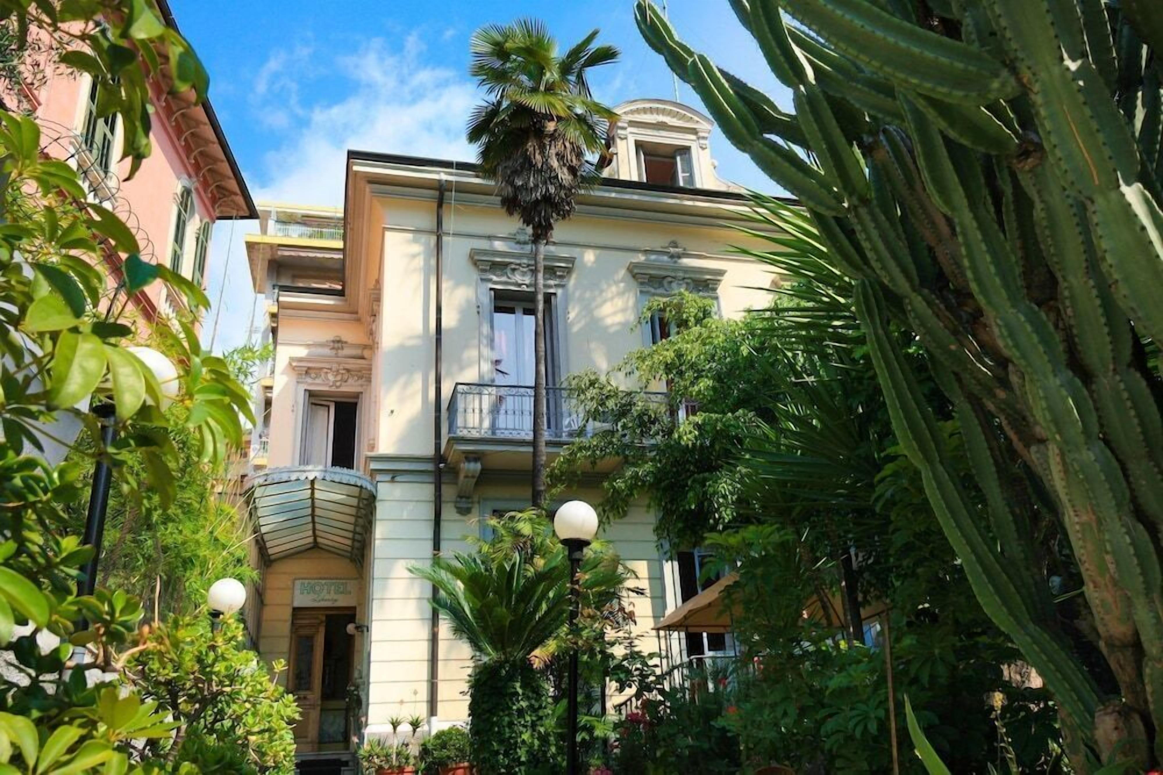 Un hotel con facciata classica, un balcone e vegetazione rigogliosa, tra cui una palma.