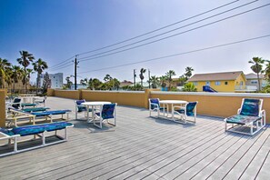 Una terraza de madera con sillones de descanso y mesas, con vistas a una playa con palmeras y un edificio amarillo al fondo.