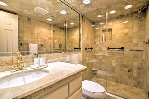Un baño con un espejo grande, un lavabo con grifo dorado, cabina de ducha con mampara de vidrio y paredes de azulejos color beige.