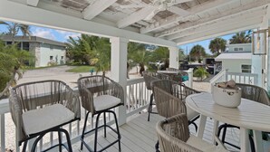 A covered outdoor seating area with wicker chairs and a table, overlooking a street with houses and palm trees. - Bluewater Beach House will not disappoint (Fort Myers Beach)