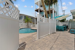A backyard with a swimming pool, a white fence, and a patio area with chairs.