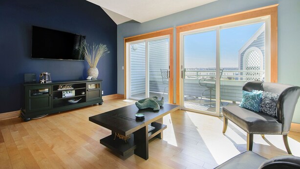 A modern living room with a dark blue wall, a flat-screen TV, a dark wooden coffee table, a gray sofa with blue and gray cushions, and a large sliding glass door leading to a balcony.
