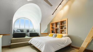 A bedroom with a large arched window, a bed with white bedding, a wooden shelf unit, and a staircase.