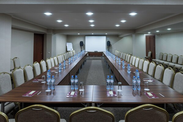 Un salón de conferencias con una larga mesa, sillas, botellas de agua y una pantalla para proyector.