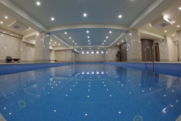 A large indoor swimming pool with clear blue tiles, surrounded by walls with a tiled pattern, and a ceiling with recessed lighting. - Astoria Tbilisi (Tbilisi)