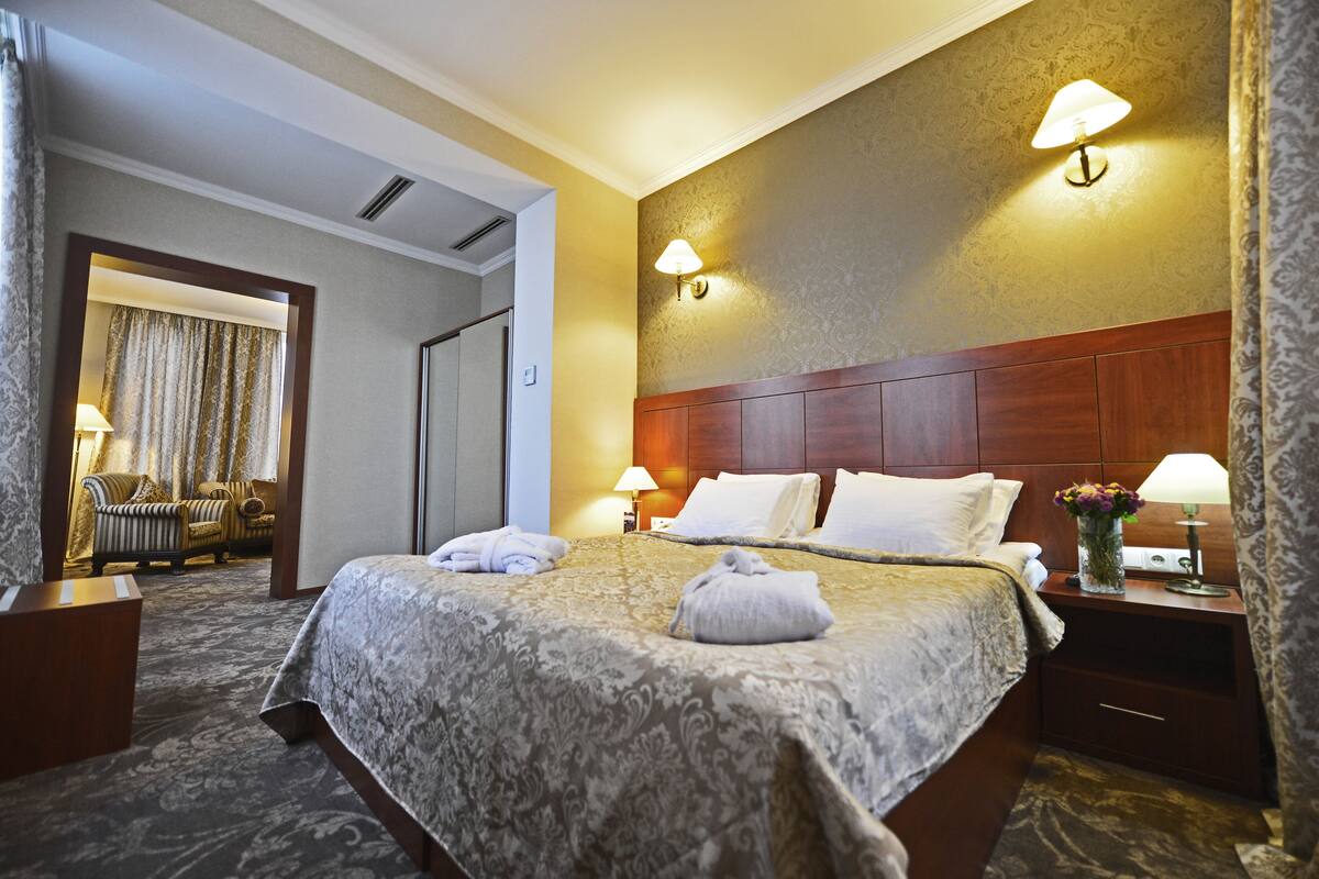 Ein Hotelzimmer mit einem großen Bett, zwei Nachttischlampen, einem Nachttisch mit Blumenvase und Blick auf einen Flur mit Tür und Sitzecke.