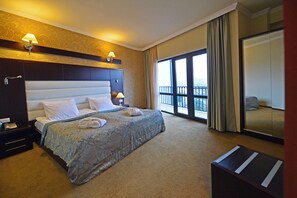 A hotel room with a large bed, two bedside lamps, a nightstand, a desk, a chair, a large window with a view, and a sliding door to a balcony. - Astoria Tbilisi (Tbilisi)