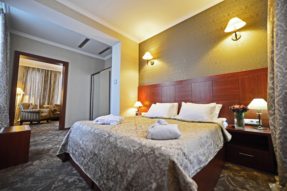 Habitación de hotel con una cama grande, dos lámparas de noche, una mesita de noche con un florero y vistas a un pasillo con una puerta y un área de estar.