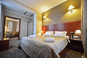 Quarto de hotel com uma cama grande, duas luminárias de cabeceira, uma mesa de cabeceira com um vaso de flores e vista para um corredor com uma porta e um espaço de estar.