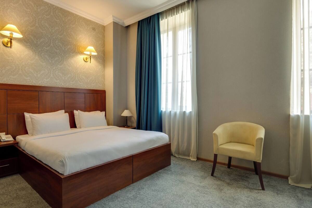 Ein Hotelzimmer mit einem hölzernen Bett, zwei Nachttischlampen, einem Sessel und einem großen Fenster mit Vorhängen.