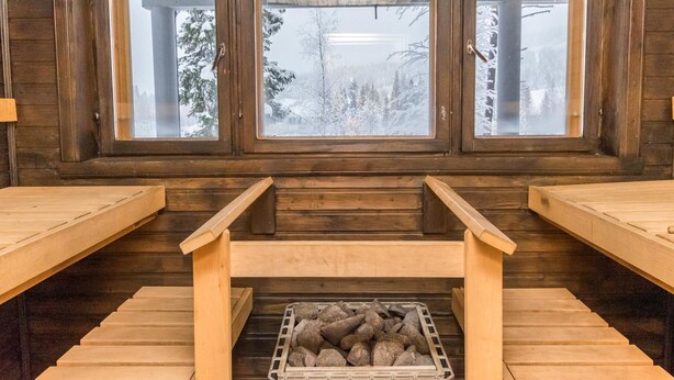 A sauna room with wooden benches, a central bench with a stone-filled container, and three large windows offering a view of a snowy forest. - Karhunpesä a by Interhome (Puolanka)