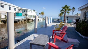 A waterfront deck with a table and red chairs, a dock with boats, and a row of houses. - Oceanside, Pet Friendly, Smart Home with-SUP, kayak with and 60 ft Dock (Marathon)