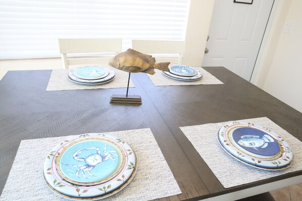 Una mesa de comedor con manteles individuales y platos con motivos marinos, un centro de mesa con forma de pez, y una puerta blanca al fondo.