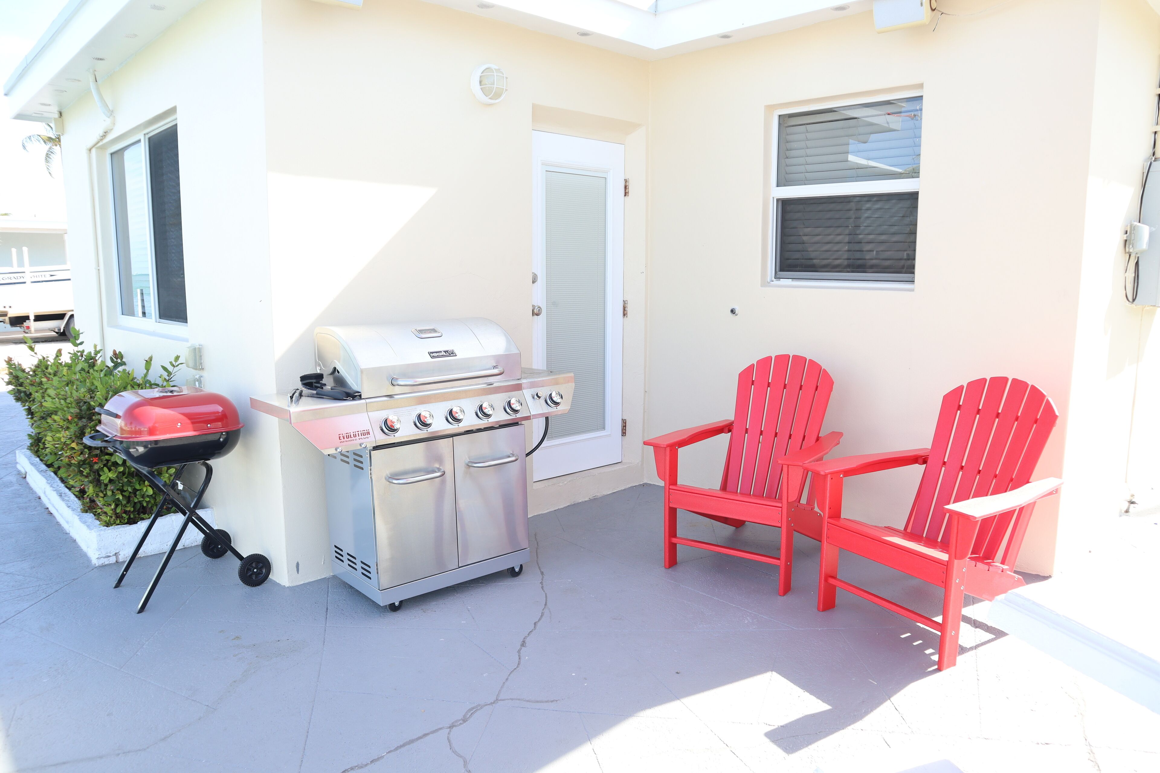 A patio with a grill, two red Adirondack chairs, and a white door.