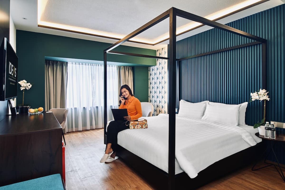 A modern hotel room with a four-poster bed, a desk with a laptop, and a small side table with a vase of flowers.