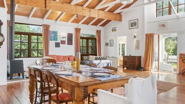A spacious dining area with a wooden table and chairs, large windows, and a high ceiling with exposed beams. - Olde Beach Lux (Waikanae Beach)
