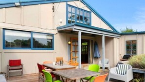A wooden deck with a table and chairs, a covered patio area, and a building with a blue roof and windows. - Olde Beach Lux (Waikanae Beach)