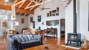 A spacious living room with a high ceiling, exposed wooden beams, a fireplace, and a cozy seating area. - Olde Beach Lux (Waikanae Beach)