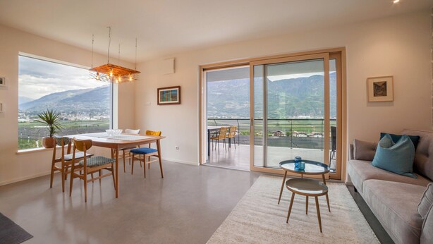 Een ruime woonkamer met een eettafel, stoelen, een bank en een balkon met uitzicht op bergen.