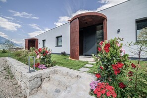 A modern building with a stone retaining wall, a wooden archway, and a garden with red and pink flowers. - Holiday Apartment "Das Eckige" with Mountain View, Wi-Fi, Garden & Terrace (Tscherms)