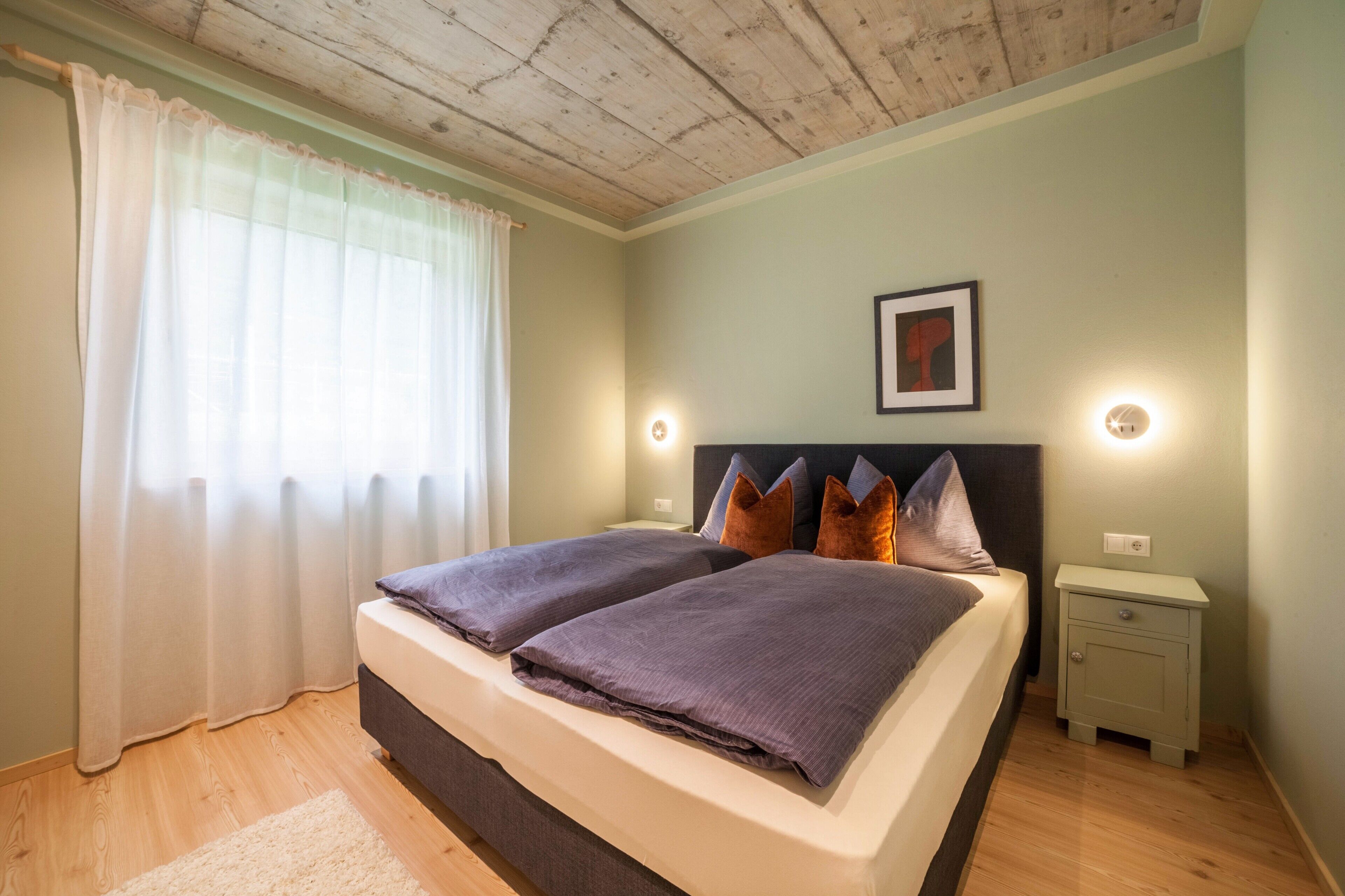 Ein Doppelbett mit Kopfteil, zwei Nachttische, ein Beistelltisch, ein gerahmtes Bild an der Wand und ein Fenster mit durchsichtigen Vorhängen.