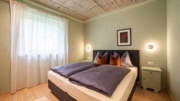 Ein Doppelbett mit Kopfteil, zwei Nachttische, ein Beistelltisch, ein gerahmtes Bild an der Wand und ein Fenster mit durchsichtigen Vorhängen.