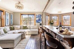 Un salon moderne avec un canapé d’angle, un coin bar avec des tabourets et une grande fenêtre donnant sur un paysage arboré.