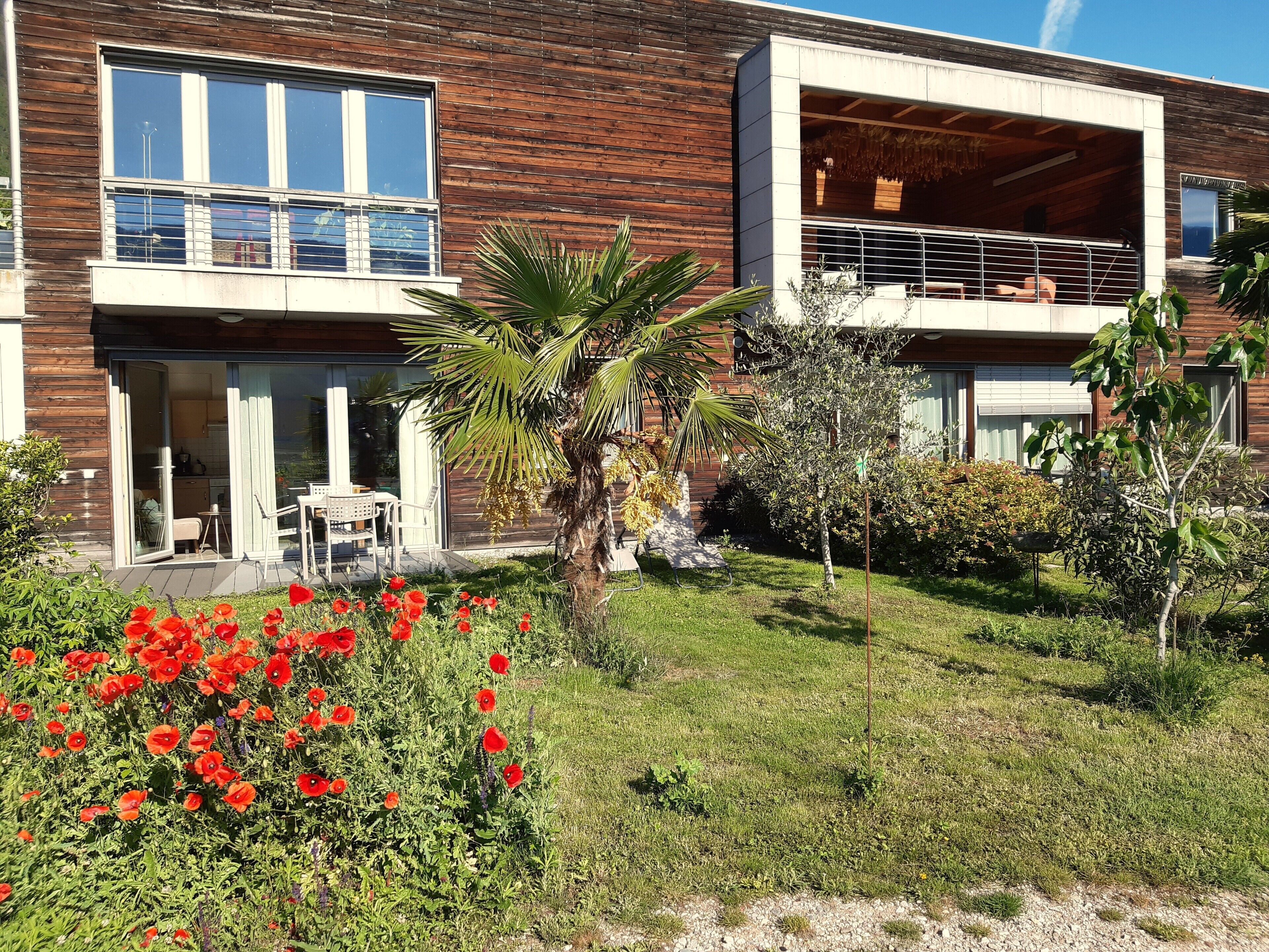 Ein modernes, zweistöckiges Haus mit einem Balkon, einer kleinen Terrasse mit Gartenmöbeln und einem Garten mit roten Blumen.
