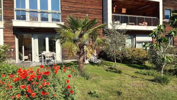 Ein modernes, zweistöckiges Haus mit einem Balkon, einer kleinen Terrasse mit Gartenmöbeln und einem Garten mit roten Blumen.