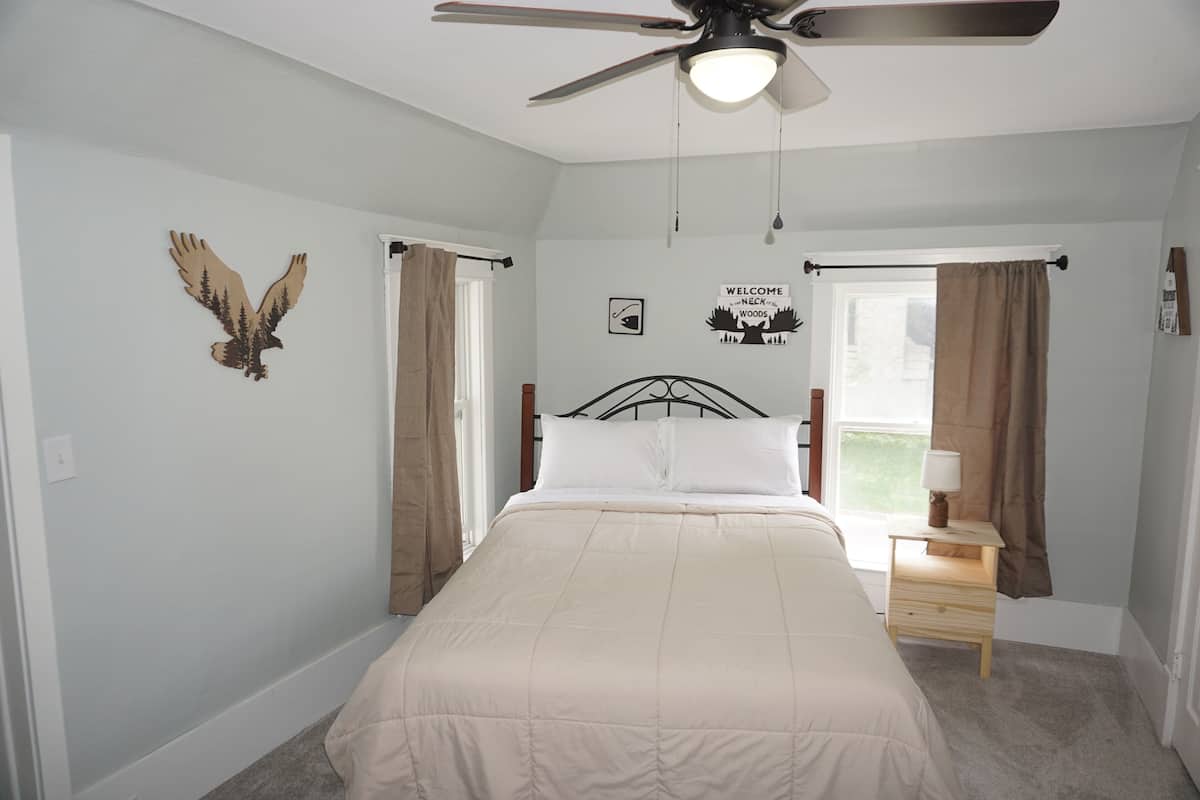 A bedroom with a bed, a ceiling fan, a nightstand, a window with curtains, and a wall decoration.