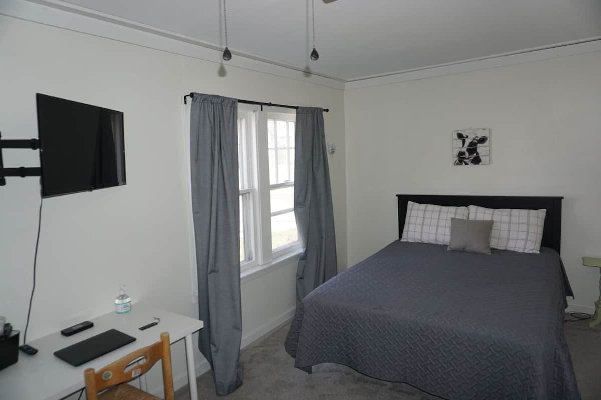 A bedroom with a bed, a desk, a chair, a TV mounted on the wall, and a window with curtains.