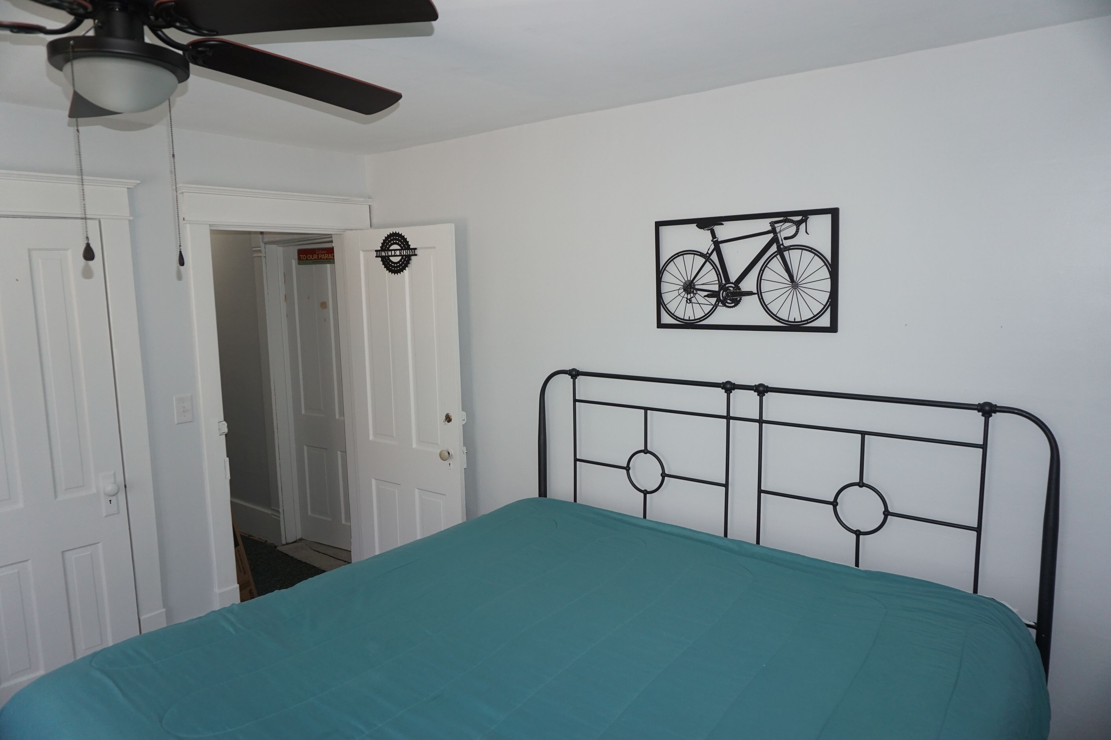 A bedroom with a teal bedspread, a metal headboard, a ceiling fan, and a wall-mounted bicycle artwork.