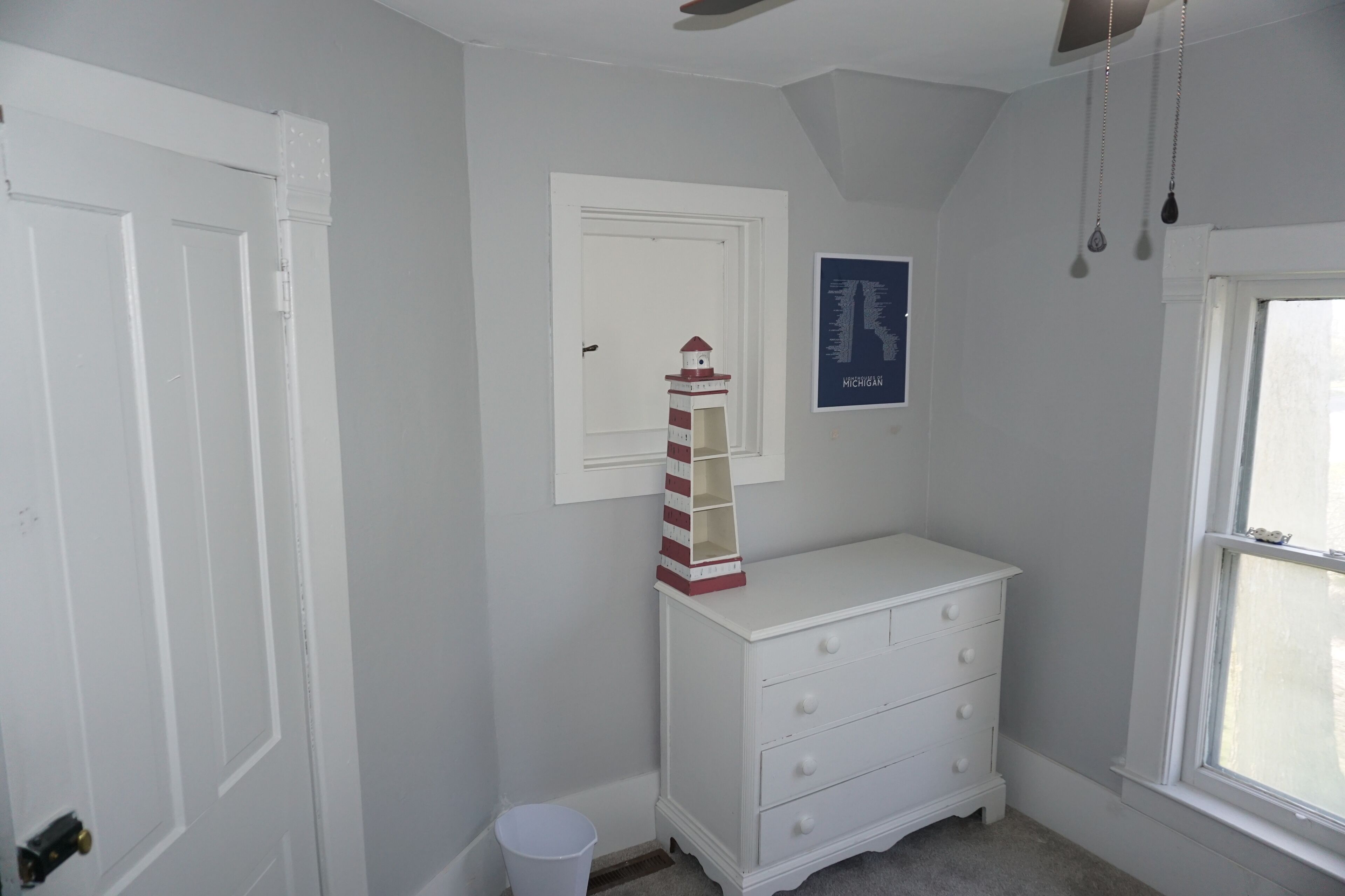 Ein Zimmer mit einem weißen Schminktisch, einer kleinen weißen Mülltonne, einer weißen Tür, einem weißen Deckenventilator und einem Fenster mit Außenblick.