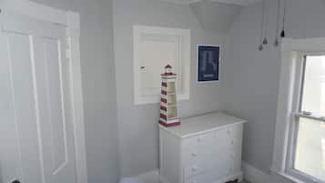 Ein Zimmer mit einem weißen Schminktisch, einer kleinen weißen Mülltonne, einer weißen Tür, einem weißen Deckenventilator und einem Fenster mit Außenblick.