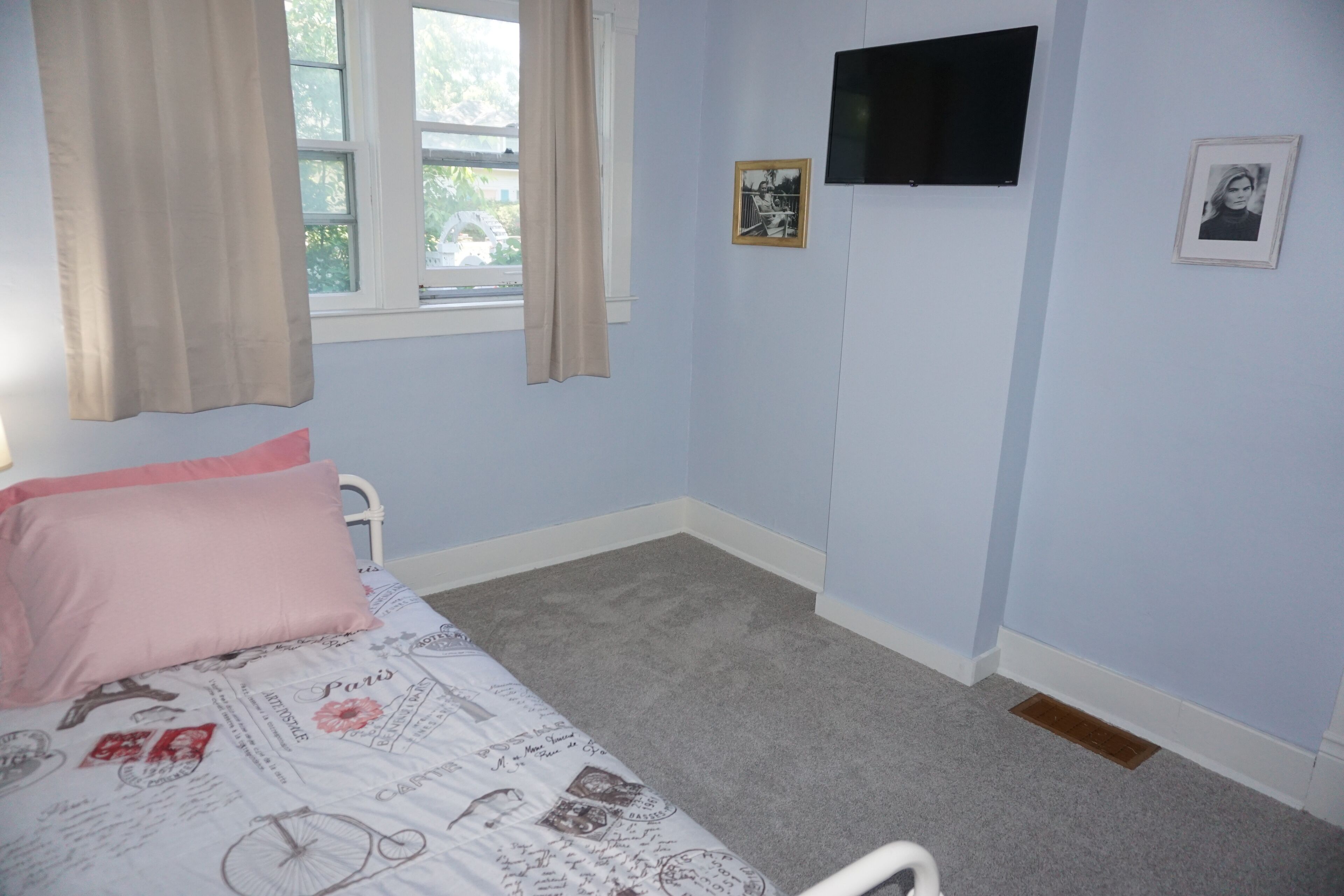 A bedroom with a bed, a window with curtains, a flat-screen TV, and two framed pictures on the wall.