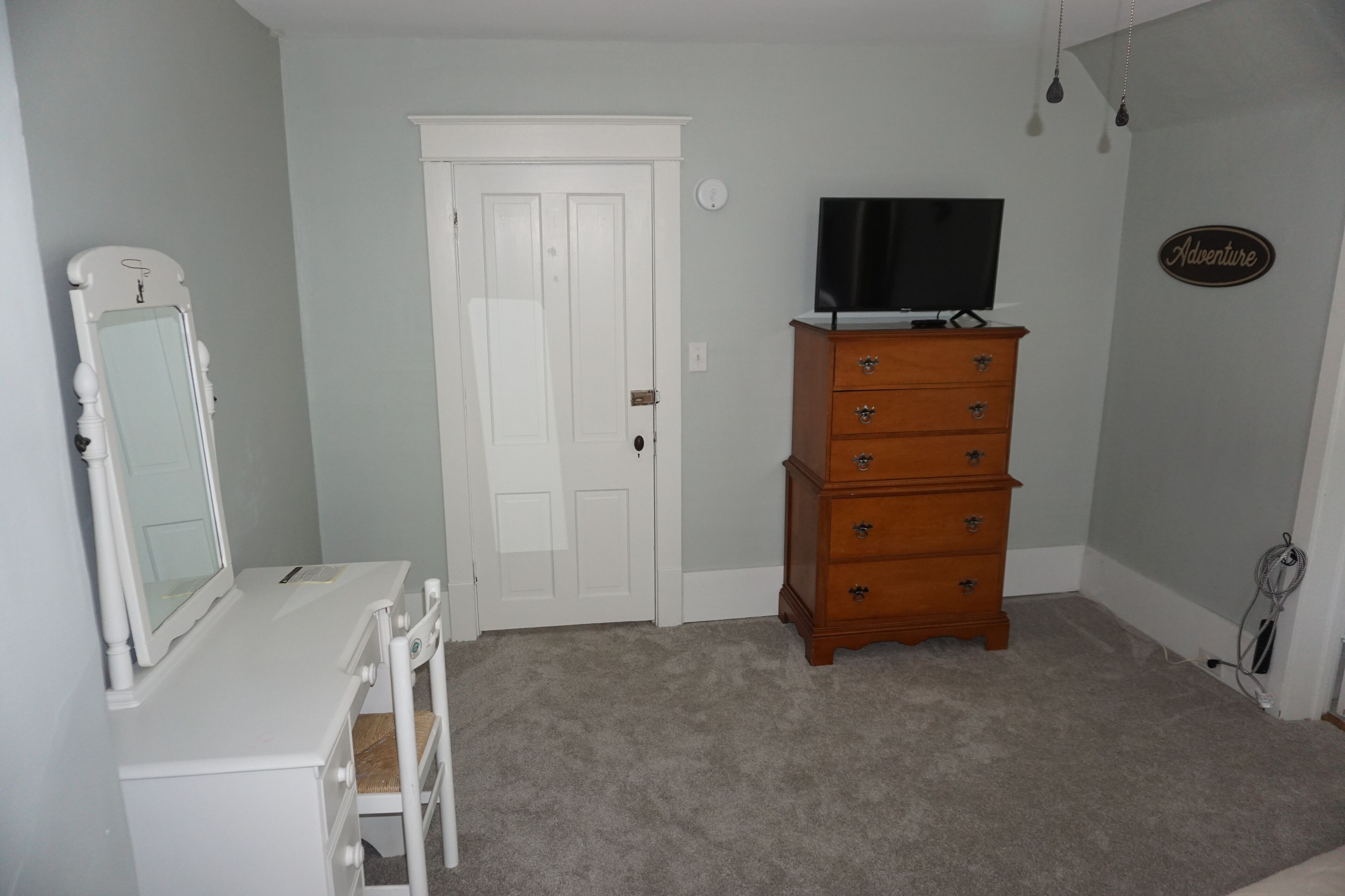 A bedroom with a white dresser, a television on top, a door, and a wall sign that says "Adventure".