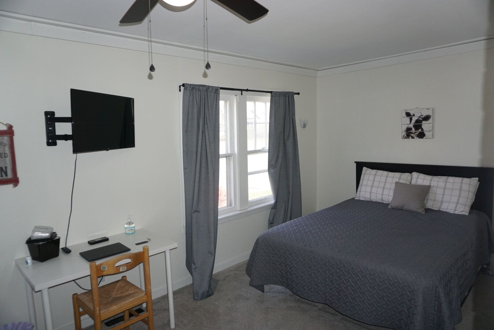 Une chambre à coucher comprenant un lit, un bureau avec une chaise, un téléviseur fixé au mur et une fenêtre avec des rideaux.