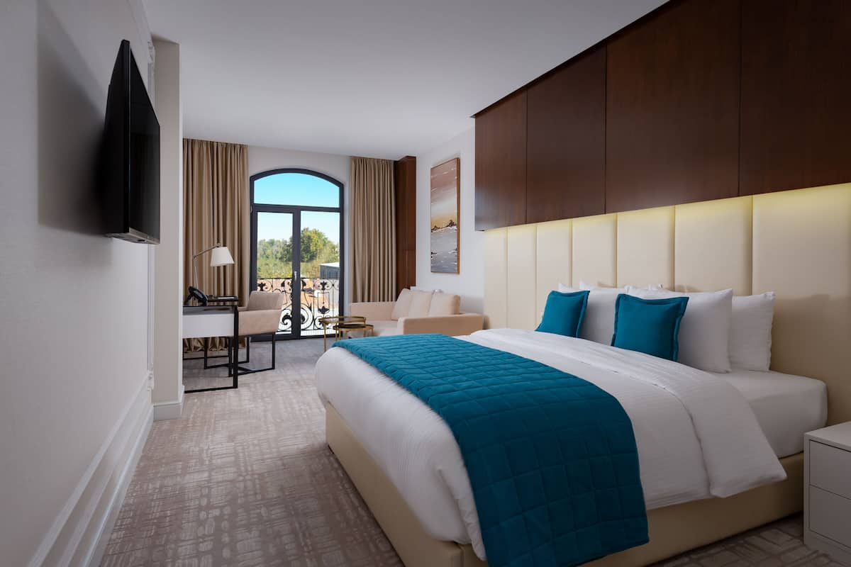 A hotel room with a large bed, a desk with a chair, a TV mounted on the wall, and a window with curtains.