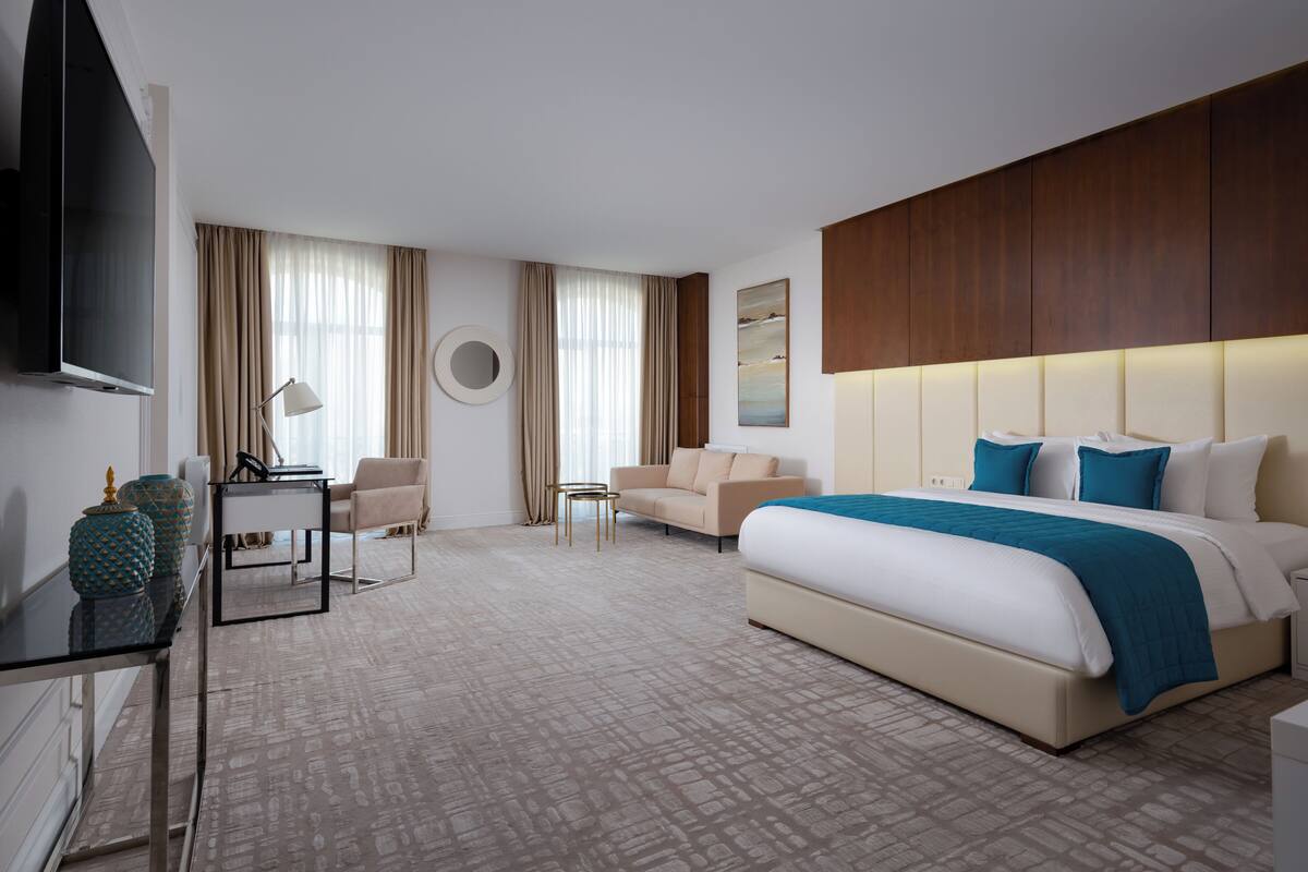 A hotel room with a large bed, a desk with a lamp, a chair, a TV, and a wall-mounted mirror.