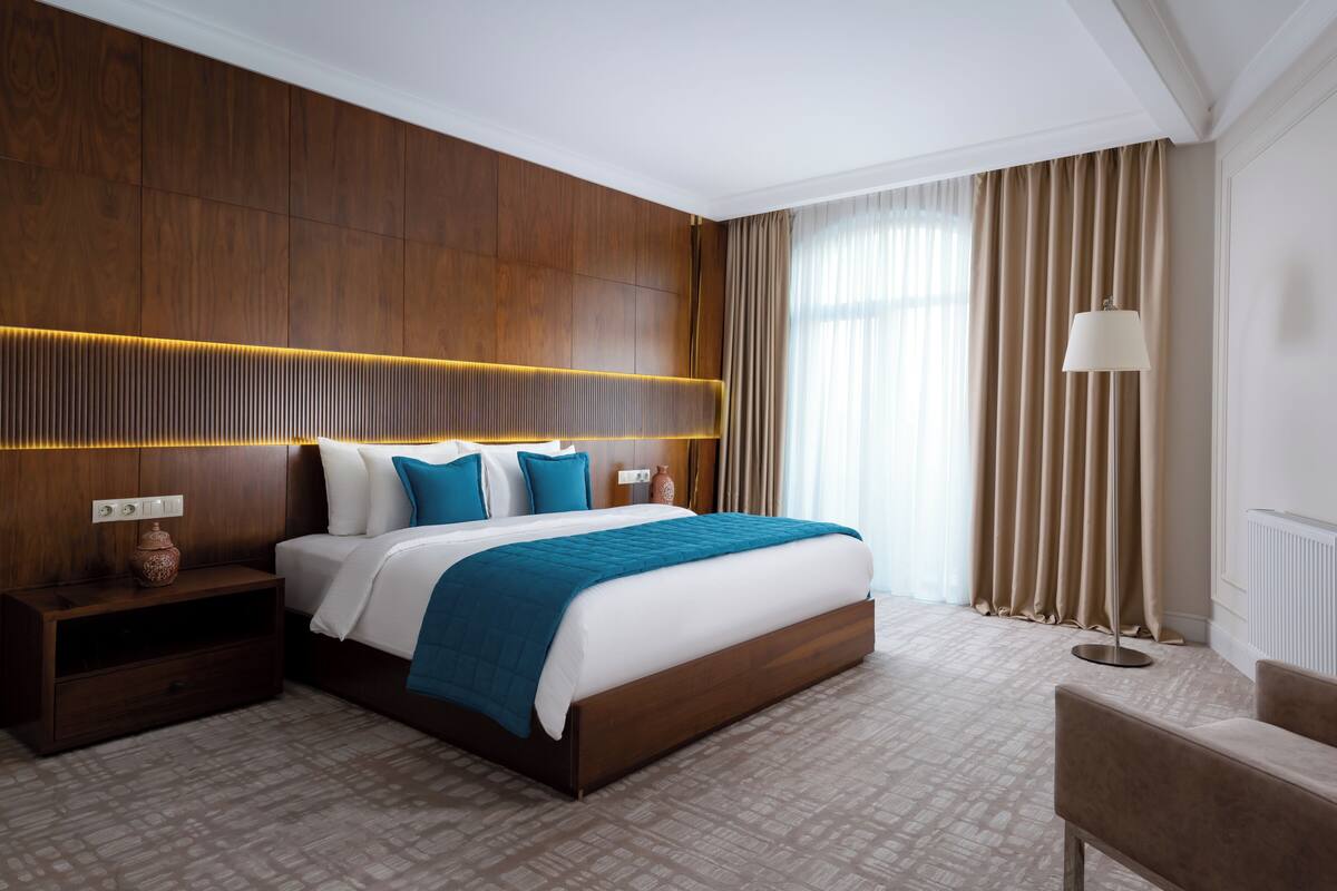A hotel room with a wooden headboard, a large bed with white and blue bedding, a nightstand with a vase, a floor lamp, and a chair.