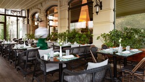 An outdoor dining area with wicker chairs and tables, potted plants, and a waiter in motion. - Mercure Tashkent South (Tashkent)