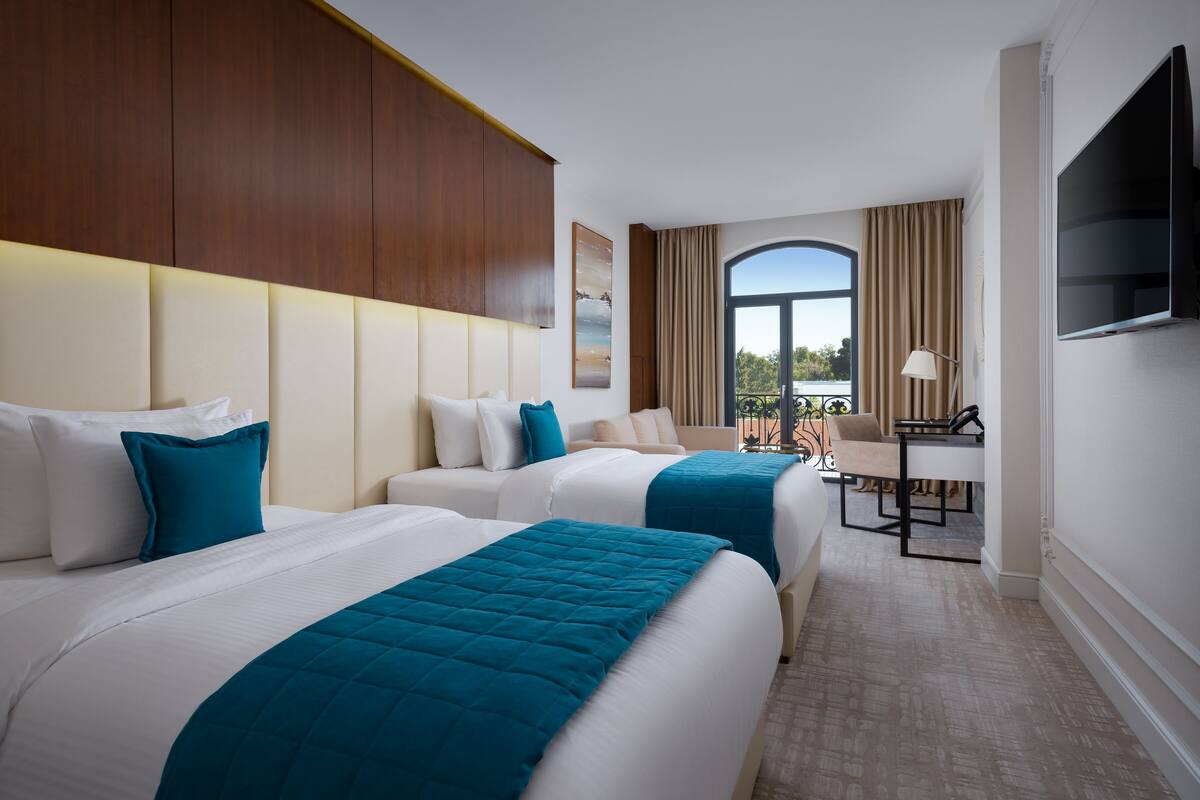 A hotel room with two beds, a desk, a TV, and a balcony with curtains.