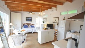 Eine kompakte Küche mit Kühlschrank, Herd und Kaffeemaschine. Es gibt einen Essbereich mit Tisch und Stühlen sowie ein Schlafzimmer mit Bett und Nachttisch.