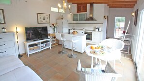 Eine moderne Küche mit weißen Schränken, ein Essbereich mit weißem Tisch und Stühlen, ein Fernseher auf einem weißen Schrank und eine Decke mit freiliegenden Holzbalken.