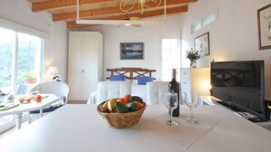 Ein Zimmer mit Esstisch, Obstkorb, zwei Weingläsern, Fernseher und Deckenventilator.