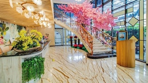 Un hall d’entrée avec un sol en marbre, un escalier orné de fleurs artificielles roses et un comptoir d’accueil agrémenté de plantes.