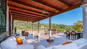 Ein überdachter Außenbereich mit weißen Sofas und orangefarbenen Kissen, einem gefliesten Boden und Blick auf eine hügelige Landschaft.