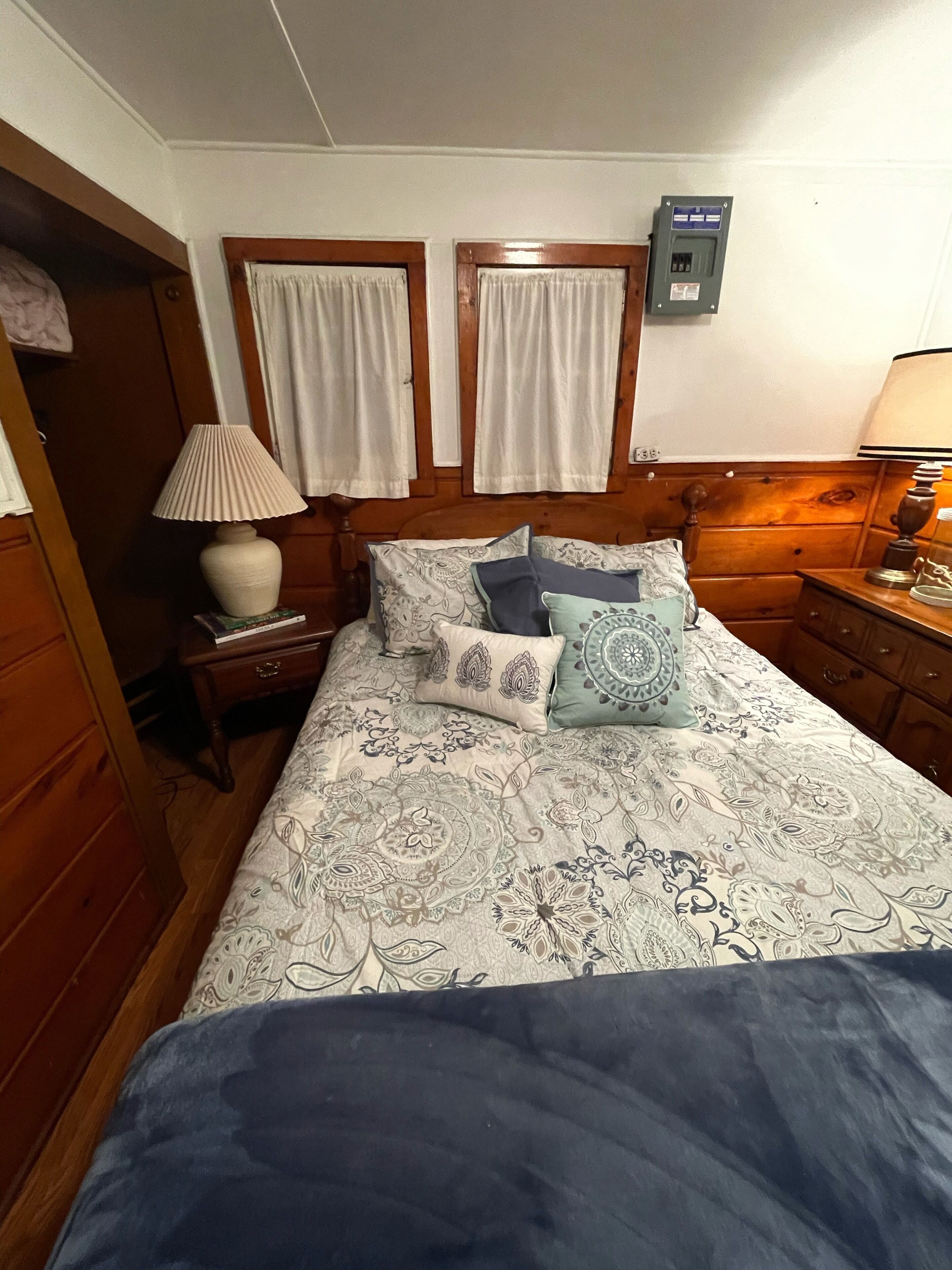 A bedroom with a wooden headboard, a bed with patterned bedding, two bedside tables with lamps, and a wall-mounted electrical box.