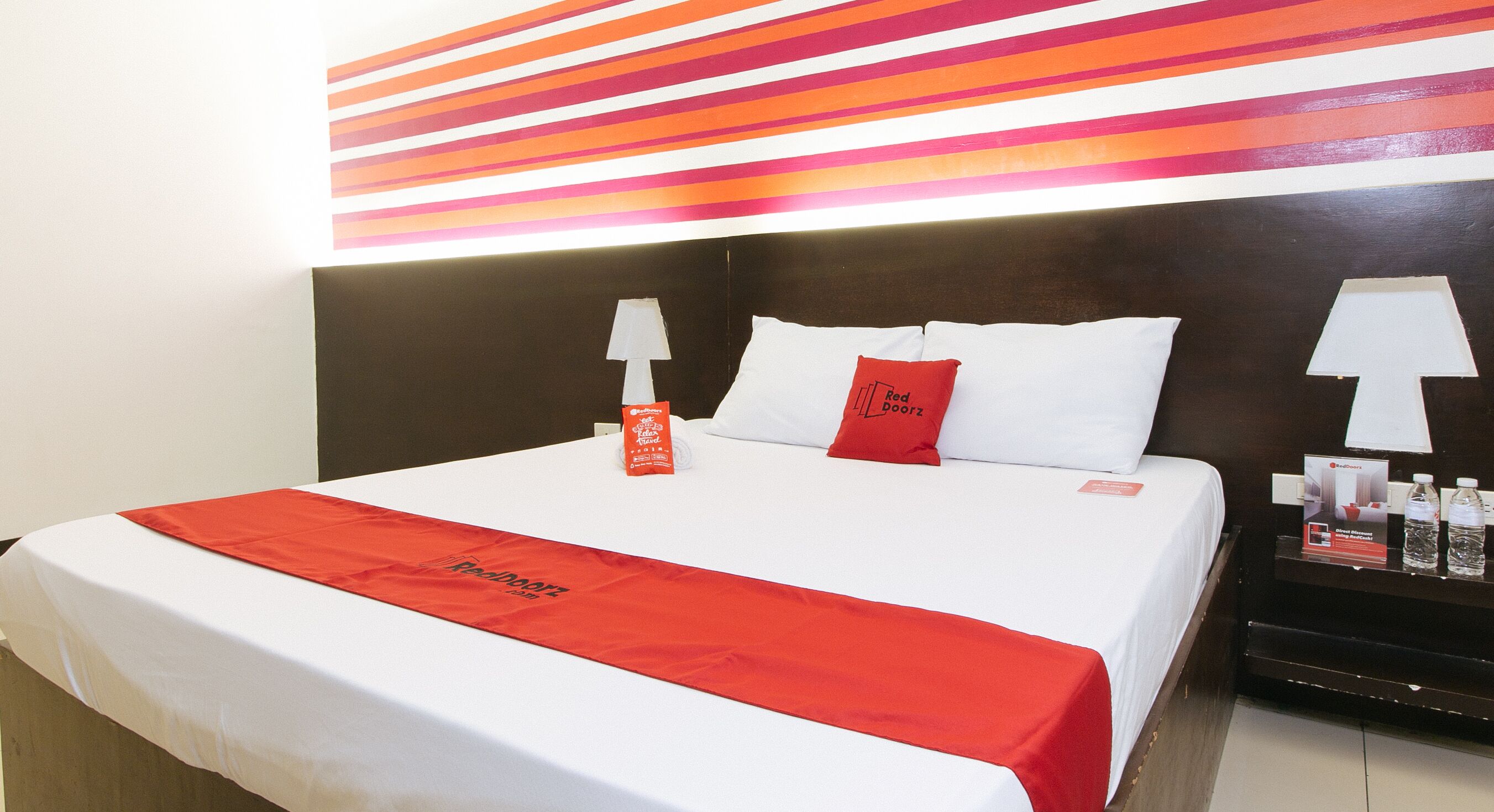 A hotel room with a neatly made bed, a red towel, and a nightstand with a lamp and bottled water.