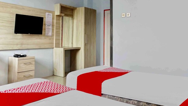 A bedroom with two beds, a TV mounted on the wall, a wooden wardrobe, and a small desk with drawers. - Super OYO 3903 Sweethome Syariah Guesthouse (Banjarbaru)